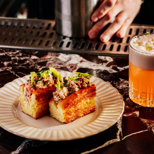 Lobster roll on plate and a cocktail sitting on a black marble bar